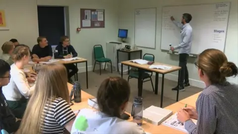 BBC A group of international students sit in a classroom during a lesson as a teacher points at a whiteboard 