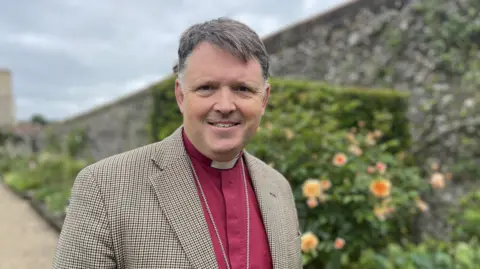 Bishop Graham in a pink clerical shirt, white dog collar and beige/brown jacket with small checks, standing in the Bishop's Garden. The background is blurred, with a flower border and a flint wall behind him.