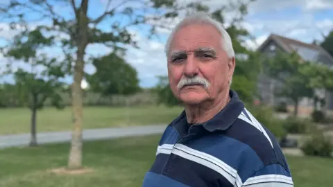 Brian is standing in a garden with lawns, trees and the edge of a barn in the background. He is wearing a blue and white striped rugby shirt and has grey hair and moustache.