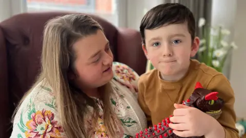 A young boy is sitting on his mother's knee. He is holding a toy dinosaur. He has dark brown hair and is wearing an orange jumper. His mother has blond hair and is wearing a floral blouse.