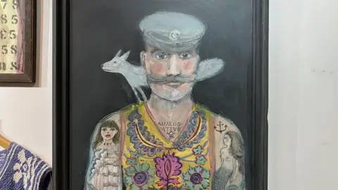 BBC/Leanne Rinne A painting of a man with a moustache wearing a sailor style hat and a yellow vest jumper with flower patterns printed on it. The man has tattoos covering his arms and a fox painted behind his shoulders. 