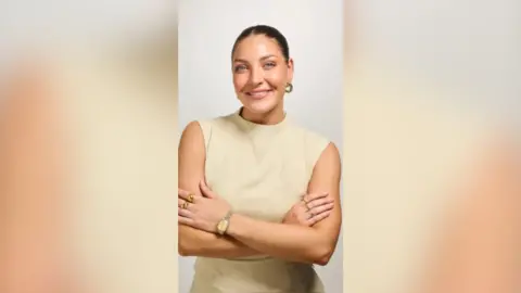 Daisy Kelly has dark, tied-back, hair and is wearing a cream dress, with her arms folded.