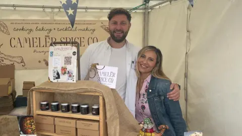 A man with a beard and brown hair with his arm around a smaller blonde woman. The man is wearing a white t-shirt and open white shirt, while the woman has a red striped shirt with floral pattern and denim jacket. They are standing behind a display of jars, boxes and banners for their spice company.