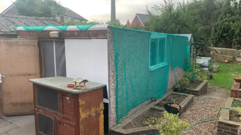 Prickly Pigs Hedgehog Rescue Outdoor garden scene featuring a long, rectangular shed or enclosed structure covered with green mesh netting along one side. A window is visible in the mesh-covered wall, and raised planting beds run alongside the structure. In the foreground is a weathered wooden hutch or storage unit with a flat top. The garden includes gravel paths, potted plants, a lawn area, and a garden hose, with trees, shrubs, and neighbouring houses visible in the background under an overcast sky.

