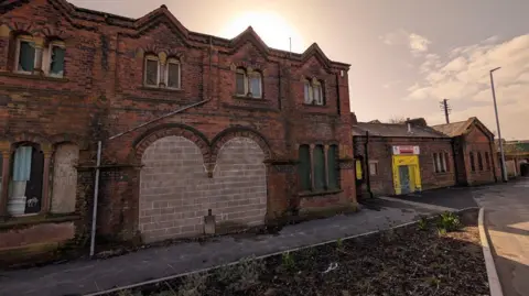 The exterior of the Strand Railway Station and club in Barrow. The building has old bricks and is boarded up. It has graffiti on some of its exterior and a red sign reading 'RAILWAYMENS CLUB' above what was once a door. The other parts of it has been boarded up.