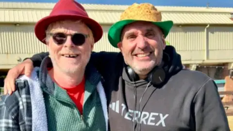 Meadows has his right arm around the artist as they pose for the photo.  He has a pair of headphones round his neck and a hat to protect him from the strong sun.  Byford wears glasses and a hat and both are grinning for the camera.
