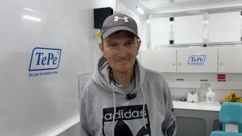 A man in a grey cap and hoodie looks at the camera. He is stood inside a clinical-looking room, with cabinets and white walls.