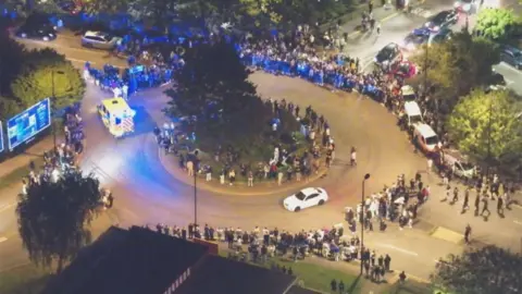 An aerial view of the car meet in question. The image has been taken a nighttime and shows a roundabout by Harbour Parade in Southampton. Crowds of people line the island in the centre of the roundabout, as well as all around the edge. A white BMW is driving around it and there are a number of vehicles parked up on the road. An ambulanc with it's blue lights is attempting to exit the roundabout, with a crowd of people in the way.