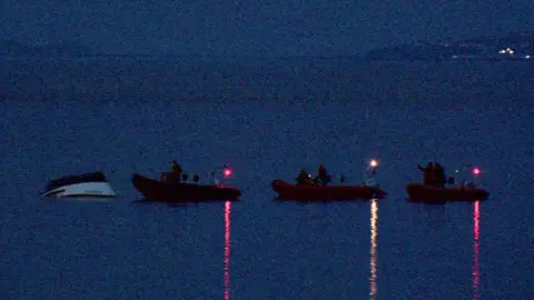 Three RNLI boats arrive towards a white and black capsized boat in a background of still blue water and sky.