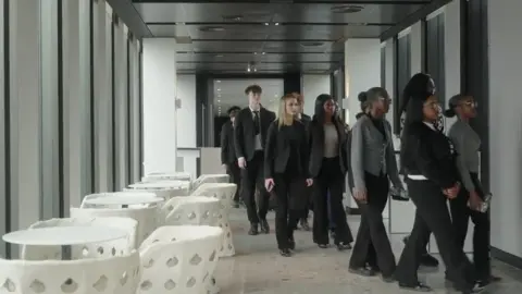 Students walk into the office which is sleek and corporate. They are dressed smartly. White tables and chairs can be seen to the left.