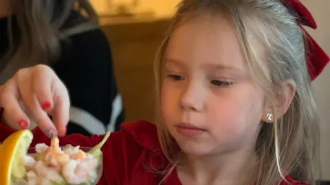 A close up of a young girl with blonde hair, Ivy, reaching to take a prawn from a prawn salad
