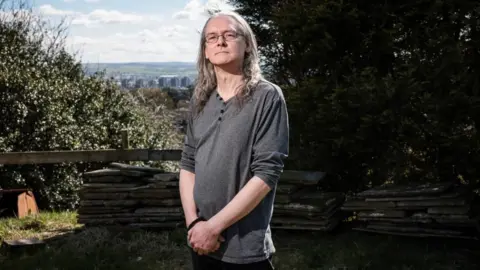 Martin Atkinson, a middle-aged man with long grey hair and glasses, stands looking serious on a grassed area. A cityscape is in the background. 