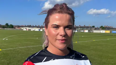 Una Simpson has long light pink hair, half tied up and is standing on a grass pitch. She is wearing a black, white a red patterned jersey t-shirt. She is also wearing a nose ring hoop.