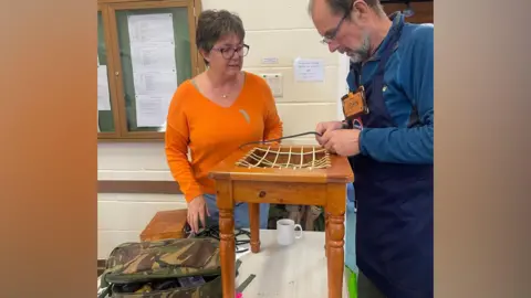 Cranleigh Repair Cafe A man with glasses, in workmen gear, repairing a small wooden table. There is a woman, in an orange jumper and jeans, watching him work.