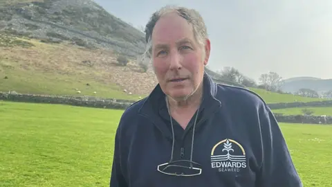 A man wearing a dark blue jumper that has a logo saying "Edwards Seaweed" over the left side of the chest looks towards the camera with a neutral expression. He has glasses on a cord around his neck. He is standing in a field with a hill behind him.