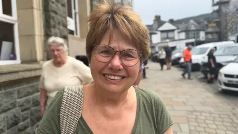 Joan, a woman with short brown hair and glasses standing on the street with a bag over her shoulder. She wears a green t-shirt. 