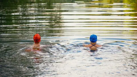 Two female open water wild swimmers in a lake are wearing orange and blue swimming caps. They are facing away from the camera and appear to be swimming breaststroke side by side with their heads out of the water. The water looks still and you can see the reflection of greenery in the distance.