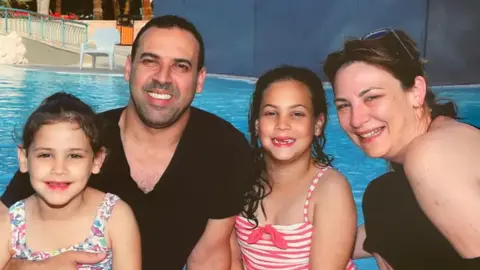 Family photo The family of four sitting in front of a swimming pool. Eli holds his younger daughter on his lap, while his other daughter sits next to him, with his wife on the right of the picture leaning in.