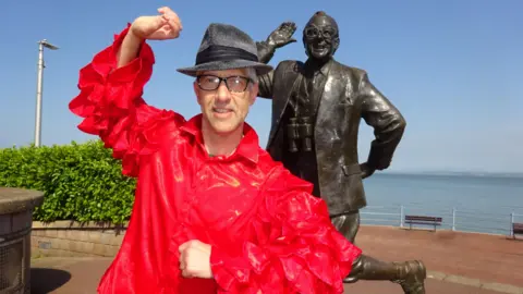 Anthony D Padgett Anthony D Padgett has a bright red PVC shirt, a trilby and spectacles and stands in front of the Eric Morecambe statue on Morecambe prom. 