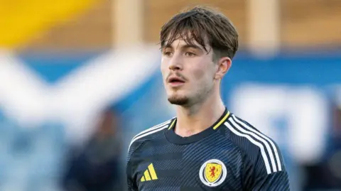 SNS A football player with dark hair and a goatee beard. He is wearing a dark blue Scotland top with white shoulder stripes and a Scotland badge. 