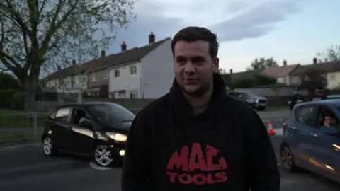 The image shows a man with brown hair. He is wearing a black hoodie. Behind him are cars and houses. 