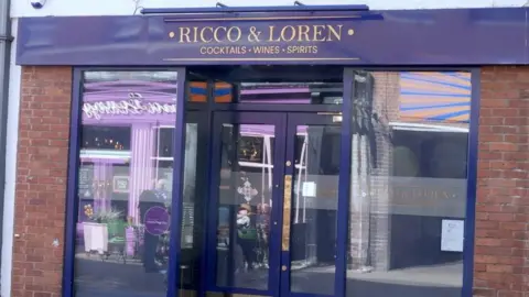 A shiny blue shop front with Ricco & Loren, cocktails wines sprits written in gold at the top of the doors.