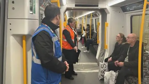 Passengers sitting on the Metro with shopping. There are some customer service staff standing next to them. The seats are linear