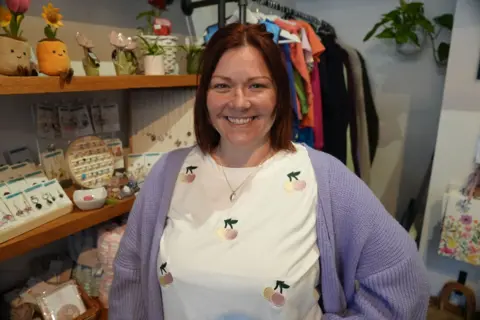 BBC news A woman in a purple cardigan and t-shirt embroidered with cherries smiles at the camera. She is standing in front of shelves holding hand made craft items. 