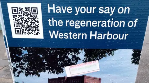 A street billboard with e QR code inviting members of the public to join the consultation. The words "Have your say on the regeneration of Western Harbour are written in white against a navy blue background.