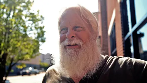 David Carter sports a full white beard - which helps him play Santa at Christmas - as he stands on a main street in downtown Kingstport