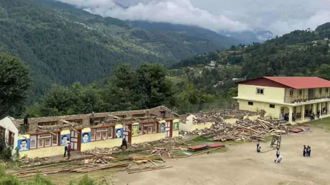 A badly-damaged school. It is at the top of a remote Nepal mountain. Mountain ranges can be seen in the background. The roof has completely come off a large section of the school and there is rubble lying scattered on the ground. There are a few people standing in the foreground.