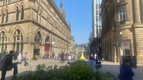 BBC/Grace Wood Market Street in Bradford on a sunny day. In the foreground people sit around a planter in the background people mill around shops and cafes