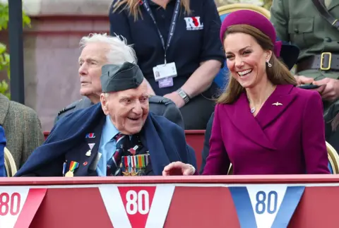 Getty Images The Princess of Wales laughs while wearing a plum-coloured dress and hat. She's sitting next to a veteran wearing military badges with bunting in front of them that says '80' on little red and blue flags
