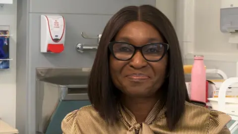 Abi Jaiyeola/BBC A woman with dark hair and a gold striped blouse with glasses.