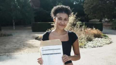A teenage girl smiles at the camera while holding a piece of paper with exam results on it. She has dark hair and is wearing a black top. She is standing outside in a school garden area.