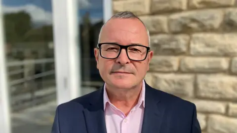 A man in glasses, a pink shirt, and a navy blazer stands in front of a brick building looking at the camera.
