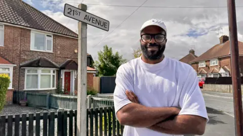 BBC A man wearing a white cap, black rimmed glasses and a white t-shirt stands with his arms folded on a street corner. He is standing in front of a road sign which reads "21st Avenue" and a number of red brick houses are visible in the background.