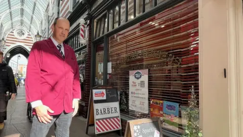 Phil Hutchinson, wearing a maroon coloured barber's jacket over a white shirt and a blue and white striped tie, is standing in a Victoria shopping arcade, next to his shop. Fixed to the exterior of the shop is a traditional red and white barber's pol. The shop itself is small and has an A Board outside advising that the shop is fully booked.  