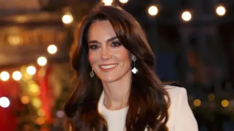 The Princess of Wales arriving for the Together At Christmas service at Westminster Abbey in 2023. She is wearing a white shift and a white blazer over it. The background is blurred, but warm Christmas lights and the outline of a Christmas tree can be seen. 