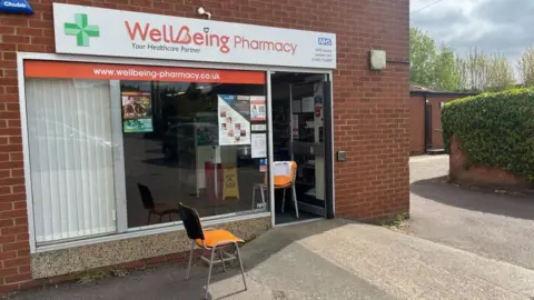 Andrew Turner/BBC The Wellbeing Pharmacy, with it's pharmacy green cross logo on the left of the signage and NHS logo at the right of that sign, above the shop window and door. There is an orange chair in the doorway, and another outside near to the access ramp to the front door. There are blinds against part of the aluminium-framed window. The building is made of red brick, and part of the boundary wall is covered in ivy, with trees growing behind the buildings and a clouded sky appearing in the upper right of the image.