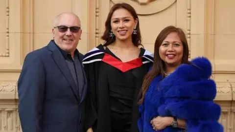 Angela Aumonier está de pie con una toga de graduación negra y roja. A su izquierda está su padrastro, David Harris, que lleva un traje azul marino. A su derecha está su madre, Angie Harris, con el pelo largo y oscuro y una chaqueta azul de pelo sintético.