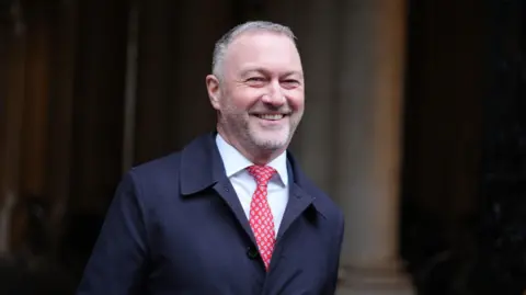 Steve Reed - a man in his 50s - smiling with short grey hair and a beard. He is walking, with the background out of focus. He is wearing a white shirt, red tie and dark blue mackintosh style coat.