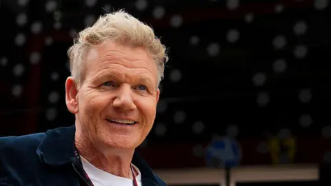 Getty Images Gordon Ramsay, a man with short blonde hair wearing a black zip-up jacket over a white t-shirt. He is smiling in front of a blurred black background.