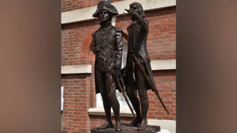 A statue features two figures both in uniform. The statue stands in front of a red brick building with an arched window