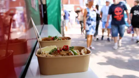 Lichfield District Council A close-up of two small cardboard containers full of food with forks sticking out of both of them. They have been placed on a shelf at the side of a trader's hut outside. People can be seen walking past on the right.