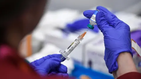 A medical staff member wearing blue gloves uses a hypodermic needle to take medicine out of a small glass vial