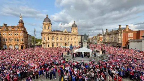 A large crowd gathers in a city square surrouded by buildings. A large statue stands on a plinth in the centre of the image.