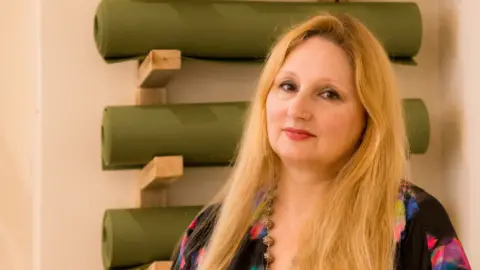 Joanne Withers Florence Achery has long blond hair and is standing in front of a rack of yoga mats.