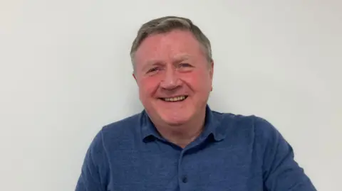 Kate Moser Andon/BBC Headshot of Dennis Jones smiling at the camera against a white wall background, with a blue polo shirt