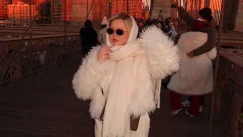 Georgia Sullivan Georgia posing on Brooklyn Bridge at sunset. She wears black sunglasses, a white faux fur coat, white scarf and white trousers. She is not looking at the camera. 